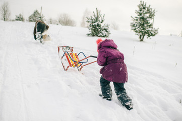 Little girl pushing sled up the snowy slide in witer day outdoors. Winter entartainment.