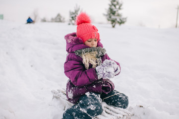 Little girl covered in snow sitting on sled with snobal in her hands in winter day. Happy child  playing with snow outdoors.