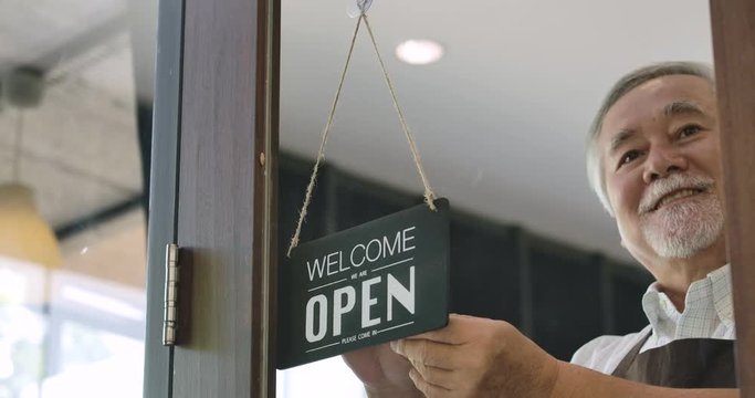 Close-up of a Senior business owner turning over a "Open" sign in the morning at her cafe window and smiling looking outside waiting for clients.