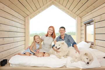 Young Caucasian family couple with baby daughter in a small modern rustic house with a large window. Lying on the bed, hugging, playing and looking out the window. Two Samoyeds. Weekend vacation.