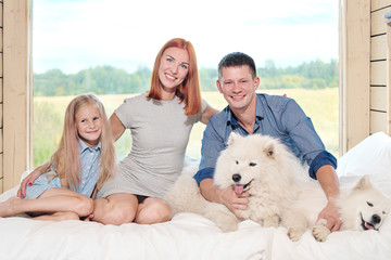 Young Caucasian family couple with baby daughter in a small modern rustic house with a large window. Lying on the bed, hugging, playing and looking out the window. Two Samoyeds. Weekend vacation.