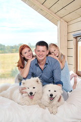 Young Caucasian family couple with baby daughter in a small modern rustic house with a large window. Lying on the bed, hugging, playing and looking out the window. Two Samoyeds. Weekend vacation.