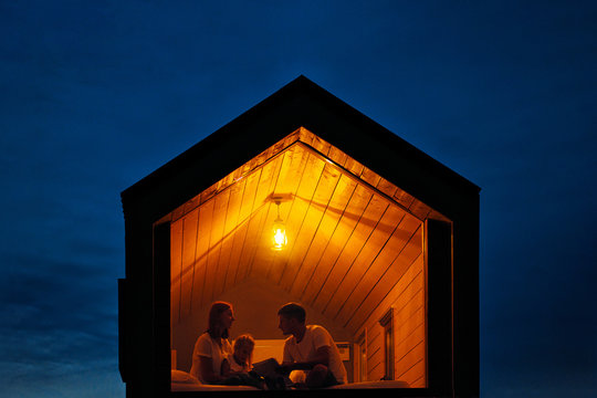 A family of three is sitting on a bed by the window, reading a book and talking. View of the outside of the house. Night blue sky. Orange warm light from an incandescent lamp.