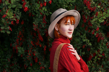 Portrait of a young attractive girl with red hair in a hat and a red sweater against the background of a bush of barberry with red berries.