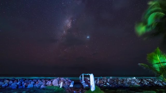 Dark night milky way time lapse with tons of stars over the ocean in Aposentillo, Nicaragua with small rock wall and palm trees in the foreground.