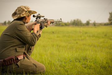 A male hunter with a gun while sitting takes aim at a forest. The concept of a successful hunt, an experienced hunter. Hunting the summer season. The hunter has a rifle and a hunting uniform. Image.