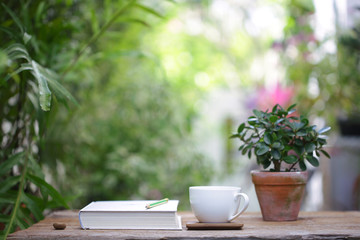 White coffee cup with small plant in old brown pot with white thick book on wooden table at outdoor with nature bokeh background