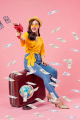 Smiling woman traveler sitting on luggage holding money gun model in holiday on pink backgrounds, relaxation concept, travel concept