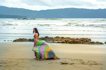 Mujer joven caminando por la orilla de playa con un manto colorido en sus manos