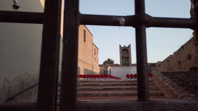 Slide shot of an auditorium with red chairs set up inside a court of a medieval castle at sunset. In the foreground an old iron gate. Camera movement from left to right.