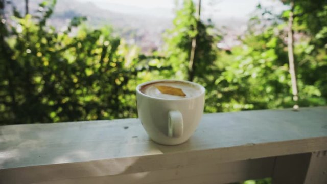 Cup of latte coffee with dry leaf inside, view of nature and city in background, dolly out, push out, zoom out.