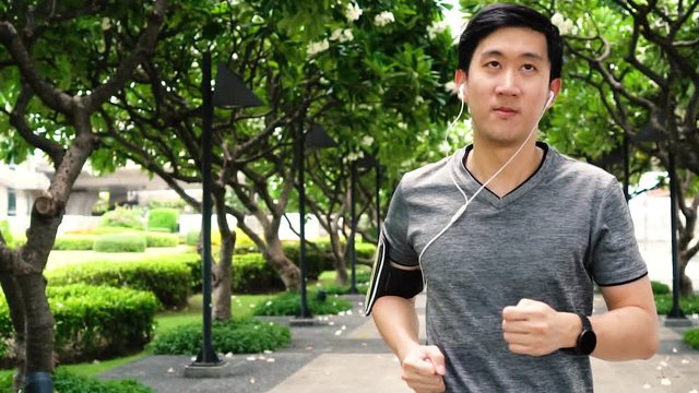 Slow motion video of young Asian man jogging while listening to music at park. Male fit runner doing exercise outdoors. 