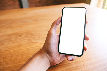 Mockup image of a hand holding black mobile phone with blank white screen on wooden table