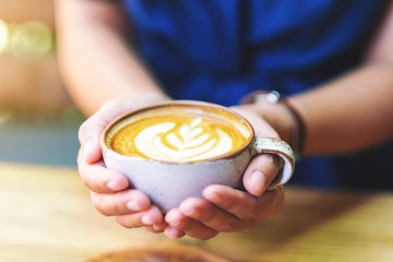 Closeup image of a woman holding a cup of hot latte coffee on wooden table