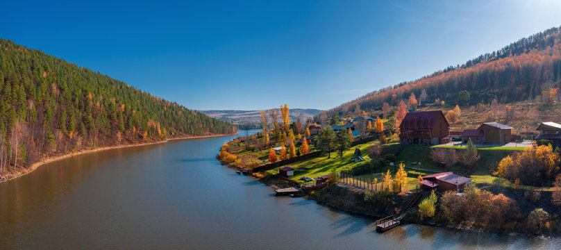 Aerial view; drone flying around little village on riverside; old buildings in autumn landscape, colorful forest; orange yellow foliage of birch trees; beautiful countryside near pond, Porogi, Ural