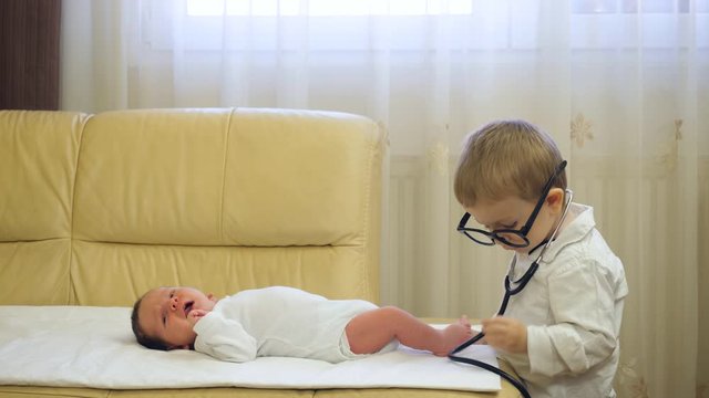 Funny little doctor child with big eyeglasses, stethoscope consult newborn baby brother