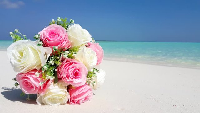 Close up shot of wedding flowers bouquet of white and pink color roses laying on the white sand beach, turquoise color sea and clear sky on background