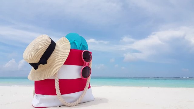 Red and white color beach bag with azure flip flops inside, pink sunglasses and flexen hat with black ribbon strip on the side laing on white sand beach 