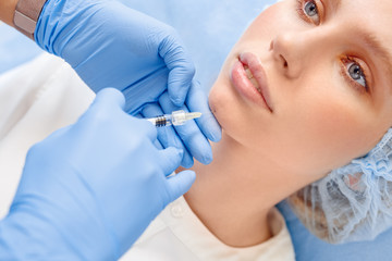 Obraz na płótnie Canvas Cosmetology Service. Young woman at beauty clinic lying while doctor in gloves doing injection of hyaluronic acid into chin face close-up