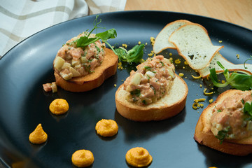 Salmon tartare on fried croutons served on a stylish black plate. Restaurant Food. Wooden bakcground. Close up