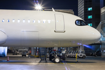 The front part of the white passenger airplane on the airport apron near the air bridge and terminal at night