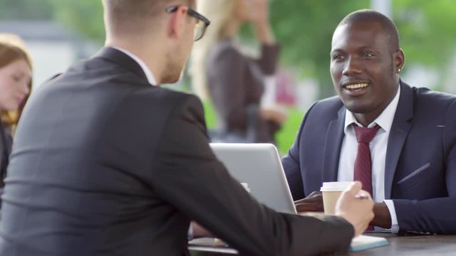 Tracking medium shot of smiling black businessman having coffee in outdoor cafe and listening to unrecognizable Caucasian colleague seen from his back