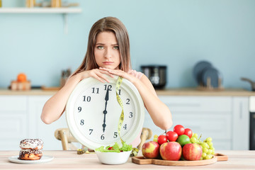 Sad woman with clock and food sitting in kitchen. Diet concept