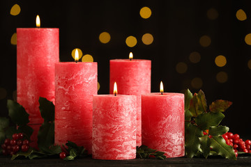Burning red candles with holly branches on table against blurred Christmas lights