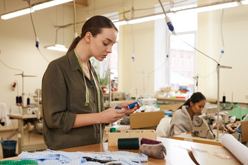 Young serious woman standing and choosing suitable thread for her dress while her colleague sewing in the background