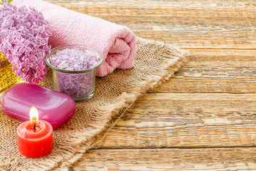 Obraz na płótnie Canvas Bottle with oil, bowl with sea salt, soap, candle and towel with lilac flowers on wooden background.