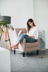 Fototapeta premium Girl decided to spend her weekend on writing novel. Young brunette in the room with white walls and daylight that comes from the window