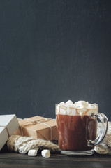 Hot cocoa with marshmallows in a glass cup surrounded by Christmas gifts and winter clothes, Christmas atmosphere and coziness