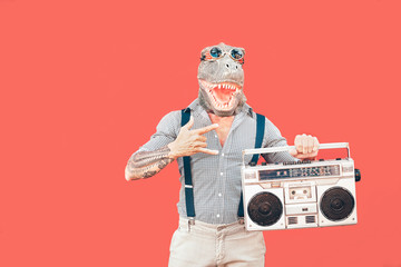 Crazy senior man wearing t-rex mask while listening to music holding vintage boombox stereo outdoor - Fashion masquerade male having fun dancing and celebrating - Absurd and funny people concept