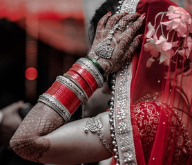 Beautiful Indian Bride in traditional Hindu wedding attire with lehnga, bridal bangles and shy pose, adjusting her netted veil with her hands.