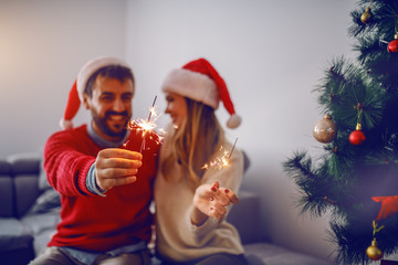 Happy handsome caucasian couple with santa hats on heads sitting on sofa in living room,celebrating New Year and holding sparklers. In foreground is christmas tree. Selective focus on sparklers.