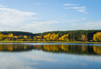Autumn park with yellow leaves and trees with a river and reflection in the water and blue sky.
