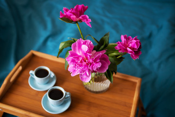 Beautiful pink peonies in a glass vase and two cups of coffee are standing on a wooden tray in bed.