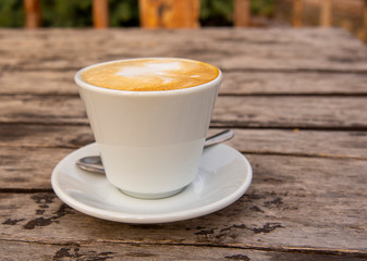 A cappuccino Cup with saucer and spoon sits on an old wooden table outside