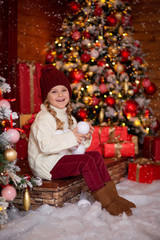 cheerful cute girl In a light knitted sweater and red hat sits on the steps of a wooden house in anticipation of the New Year holiday, in her hands she holds snowballs and has fun