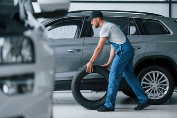 New one. Mechanic holding a tire at the repair garage. Replacement of winter and summer tires - obrazy, fototapety, plakaty