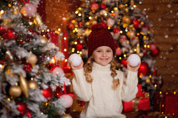 Fototapeta na wymiar cheerful cute girl In a light knitted sweater and red hat sits on the steps of a wooden house in anticipation of the New Year holiday, in her hands she holds snowballs and has fun