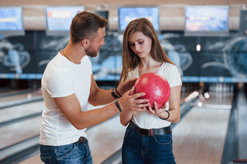 Man teaching girl how to holds ball and play bowling in the club