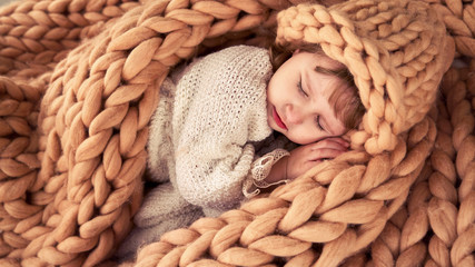 child in a hat under a blanket of natural sheep wool. Merino plaid knit covers little girl....