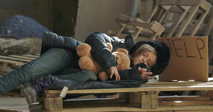 Homeless Caucasian girl in winter clothes waking up with the teddy bear on the construction site. Exhausted child living on the streets.