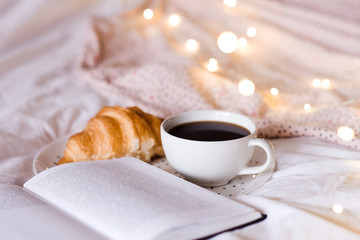 Fresh croissant with cup of hot coffee and open book in bed over Christmas lights closeup. Good...