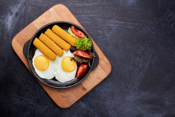 Fried fish sticks or breaded fish fillet in a cast-iron pan, close-up. Fried eggs, fresh tomatoes and salad.