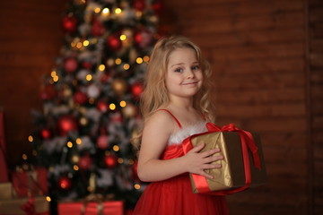 Obraz na płótnie Canvas Beautiful blonde girl in a bright Red festive dress holds a gift in her hands in anticipation of the New Year against the background of a wooden house.