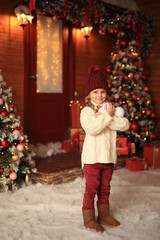 cute beautiful girl in a white sweater and red hat having fun and playing snowballs in winter on the street against the background of a wooden house. nearby are Christmas trees with lights