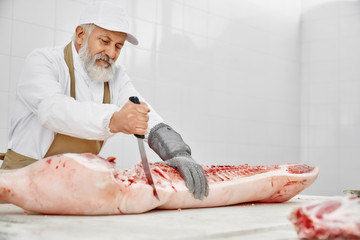 Elderly butcher in uniform cutting pork carcass with knife.