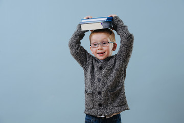 Cute little ginger boy in glasses holding pile of books on his head over blue